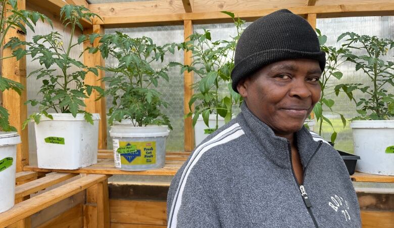 A woman in a grey sweater stands in front of a row of plants in a greenhouse.