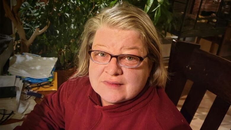 A woman sits on a chair wearing a red sweater and glasses.