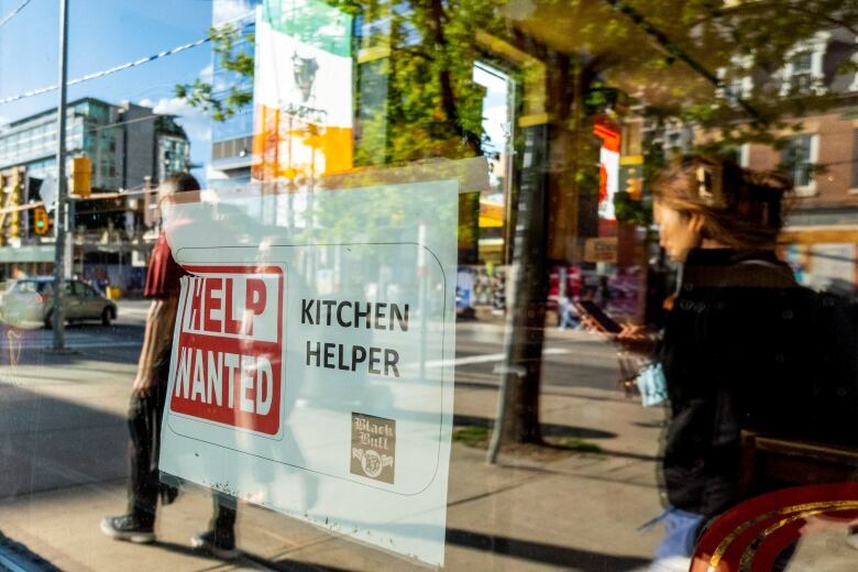 People walk past a help wanted sign adhered to a storefront window.