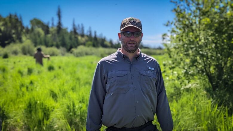 Man wearing baseball cap and sunglasses standing in grassy area.