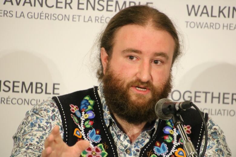 A man speaks at a microphone wearing a beaded vest.