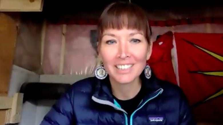 A woman smiles at the camera on a video call from inside a woodframe structure. 