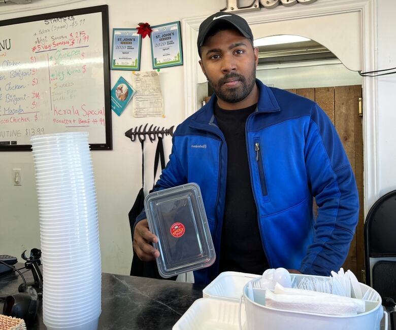 A young man is looking into the camera, holding up a plastic takeout container. More plastic containers and cutlery are piled up on the counter in front of him.