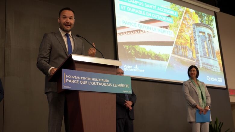 A man speaks at a lectern with a projector screen beside him.