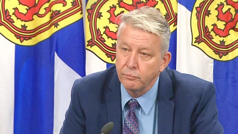 A man in a dark blue suit, light blue shirt and dark blue tie sits in front of a Nova Scotia flag.