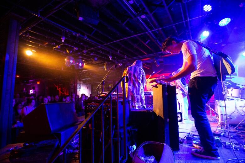 A man in a white shier plays a musical instrument on stage as a crowd looks on. The area is covered in coloured lights