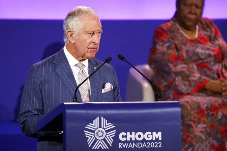 A man delivers a speech from a podium while a woman sits in the background.