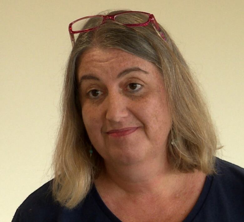 A middle-aged woman stands in front of a beige background. She has straight blonde hair down to her shoulders, and has red glasses sitting on her head.