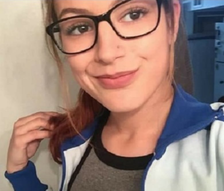 A woman with black glasses is seen wearing a grey shirt and a blue and white jacket. She is wearing a ponytail with brown-red hair and smiling at the camera.