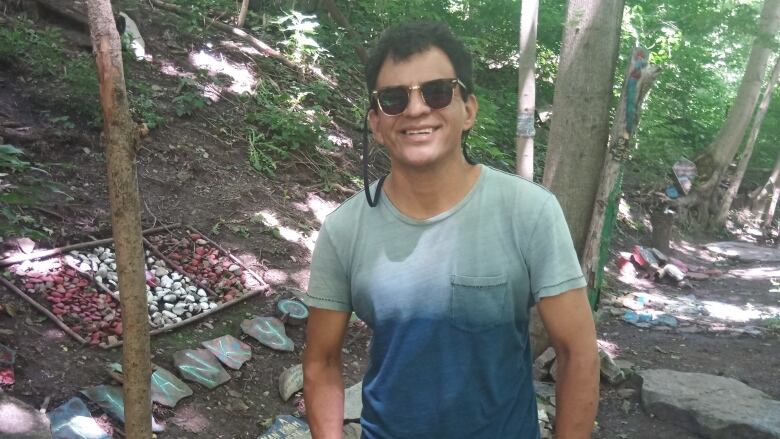 A man in sunglasses stands smiling in the forest. 