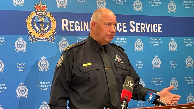 Regina Police Chief Evan Bray speaks at a podium with a blue backdrop behind him. The backdrop is covered in the Regina Police Service logo and the organization's motto: 