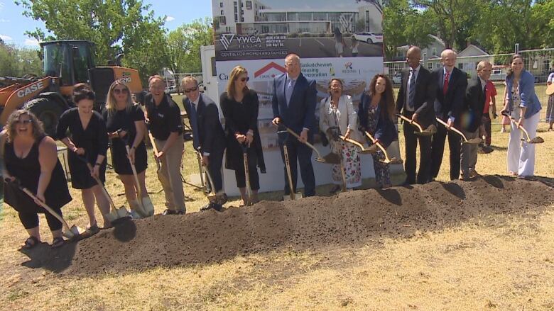 A row of 12 people holding gold-coloured shovels dig into a pile of dirt.