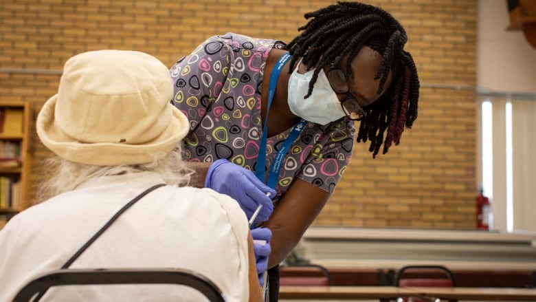 A nurse wearing a blue medical mask and latex gloves administers a vaccine to the right arm of a woman with grey hair, wearing a hat.