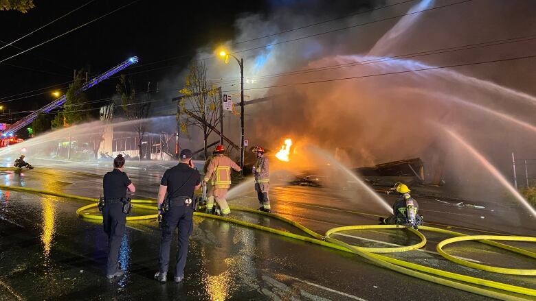 Streams of water are seen spewing from pipes, and firefighters mill around, with a fire in the background and blue warning lights.