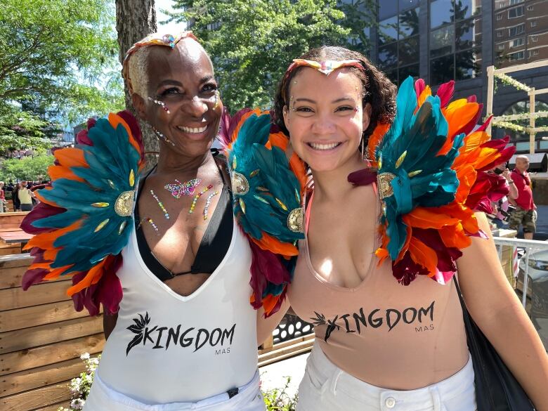 A mother and daughter stand at a parade, decked out in bright feathers and jewels around their shoulders. 