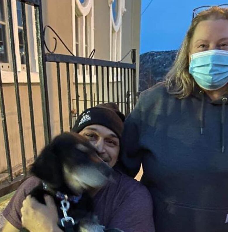 A man grins while holding a black dog in his arms. A woman has her arm around Clarke. They are outside near a residential building in St. John's. 