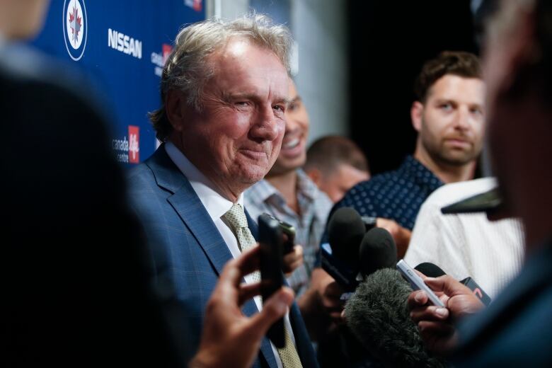 A smiling man in a suit is surrounded by reporters holding microphones.