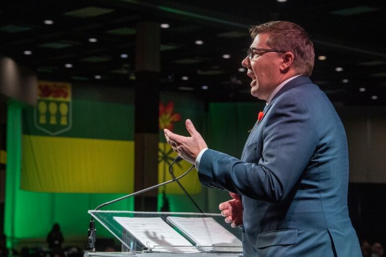 A man in a suit stands at a podium, talking to an audience.