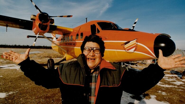 A late Billy Diamond, former Cree Grand Chief, stands with his arms outstretched smiling in front of a twin-otter plane painted bright orange.