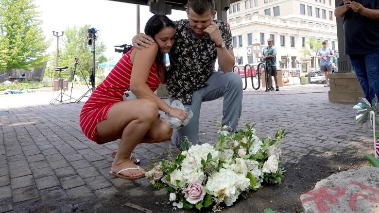 A man and woman embrace while crouching near a bouquet of flowers on the ground. 