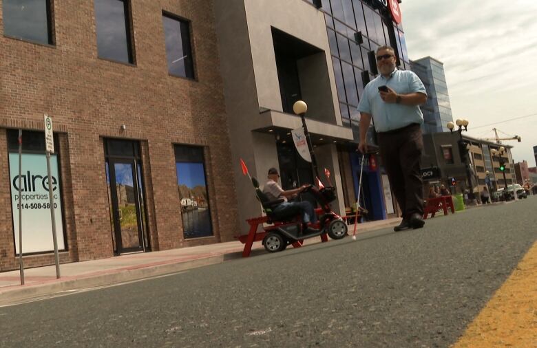 A man walks down the road in Downtown St. John's. He's blind, and is walking with a white cane and a phone to give him directions.