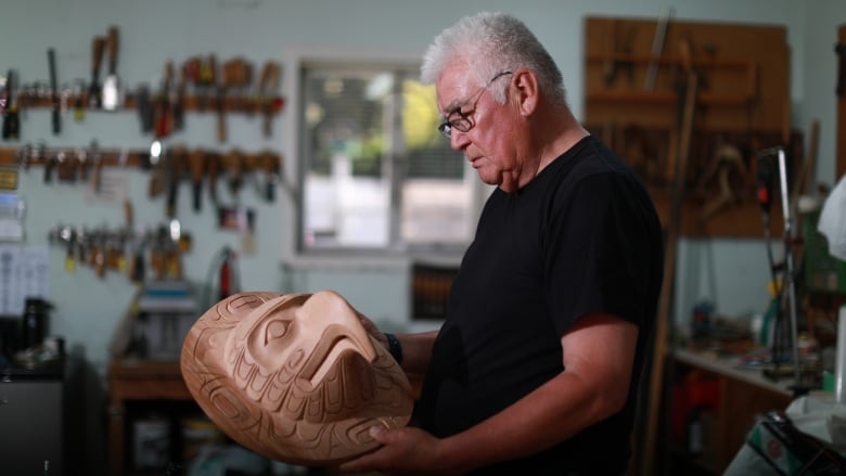 A carver, standing in his workshop, holds one of his works - a mask of a bird - in his hands. 