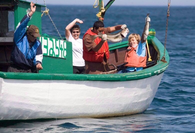 Four people wave from aboard a small fishing boat at sea. One of them is holding a large cod. 