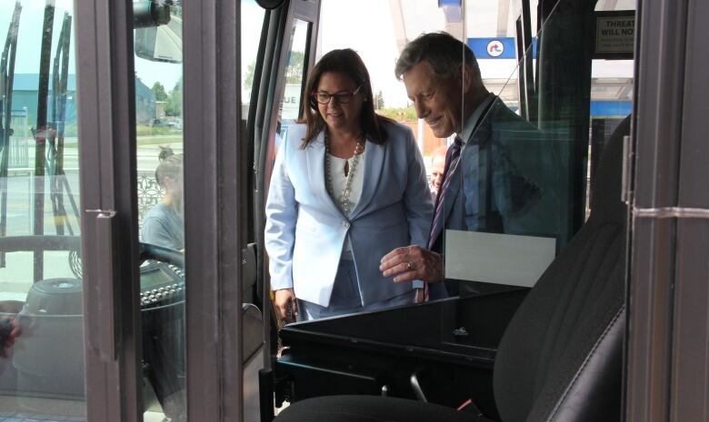 A woman and a man stand in the entrance area of a bus, looking at the driver's area of the vehicle.