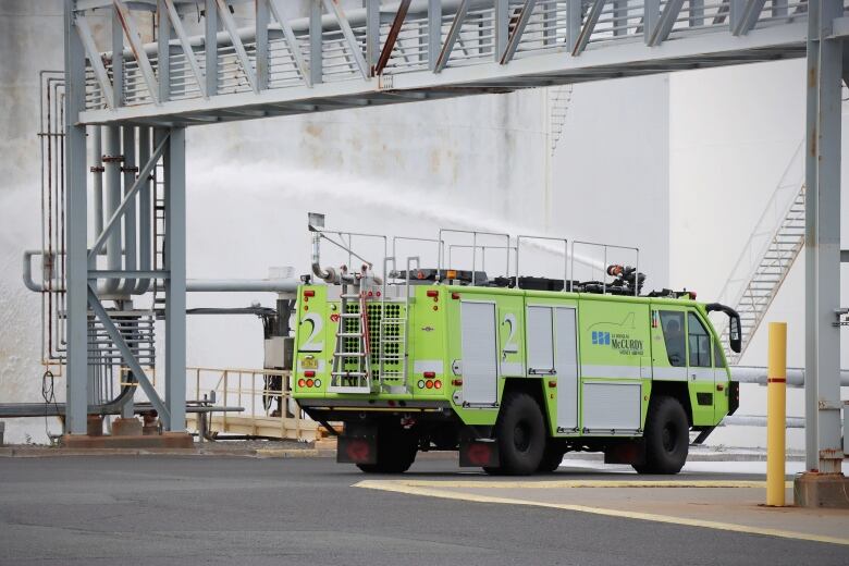 A green box-shaped fire truck sprays foam from a nozzle mounted on top in amongst large fuels storage tanks.