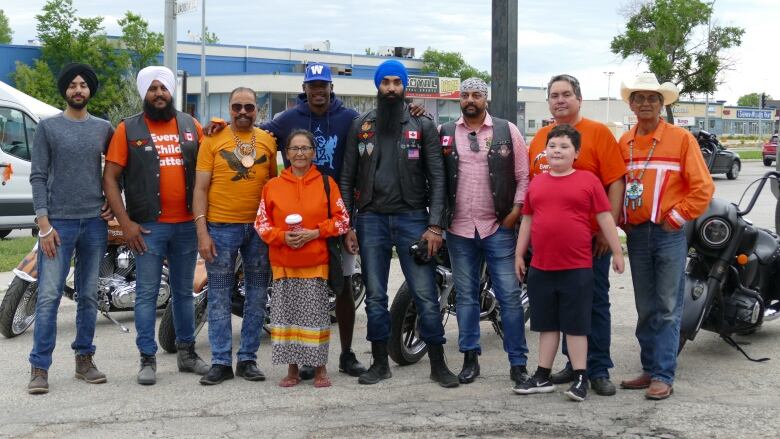 A group of 10 people, young and old, pose in a line in front of four parked motorcycles. Some people are wearing a mix of motorcycle riding-type clothing, like denim and leather vests or jackets, and some are wearing orange shirts or hoodies.