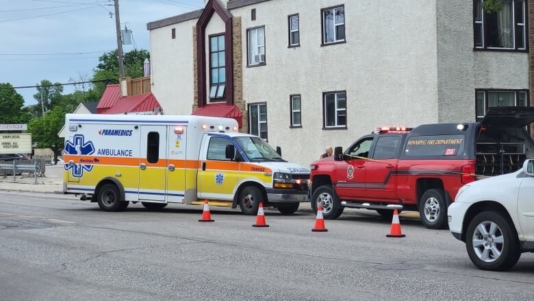 An ambulance and a vehicle with the Winnipeg Fire Paramedic Service logo are parked in front of a two-storey apartment building with small pylons on the road in front of them.