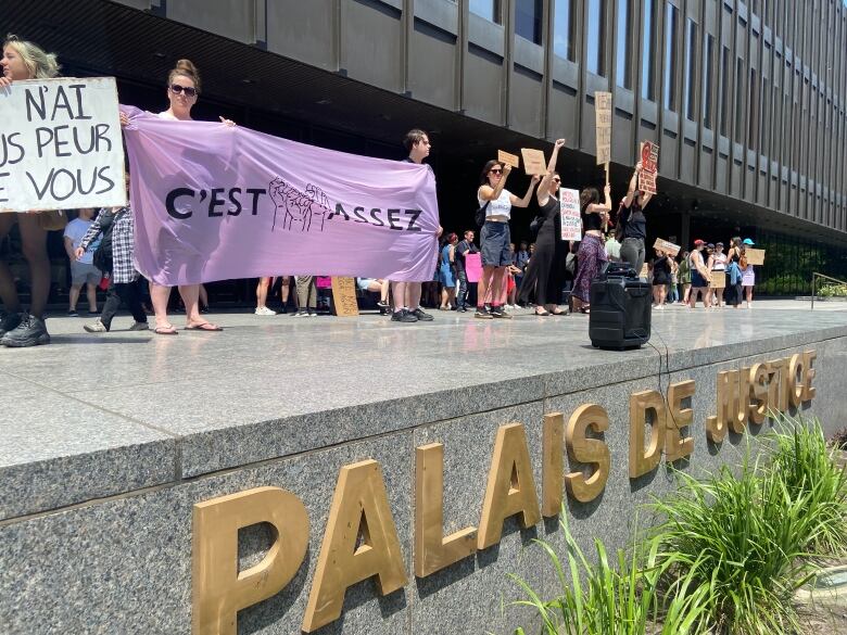 A group of dozens of protestors, many women, gather outside a courthouse with a sign reading Cest assez, French for Its enough.