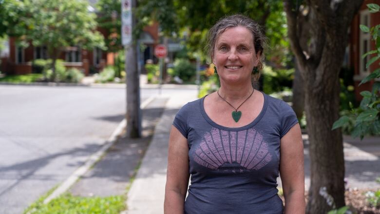 A woman wearing a T-shirt stands on a sidewalk and smiles at the camera.