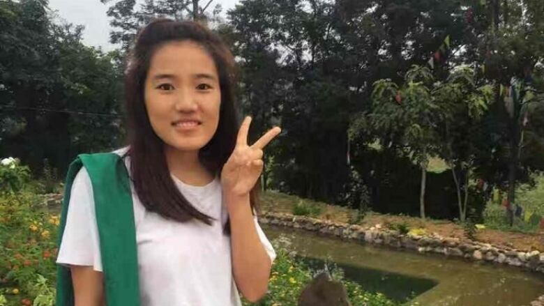 A young woman poses with a peace sign in a garden wearing a T shirt and jeans while carrying a bag