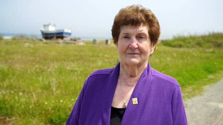 A woman wearing a purple jacket stands in front a field, which has a raised boat sitting on the land. 