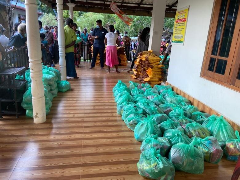 Many green plastic grocery bags filled with items lined up on a the wood floor of a porch, near piles of yellow and brown rice bags. Several people are lined up behind a fence surrounding the porch. 