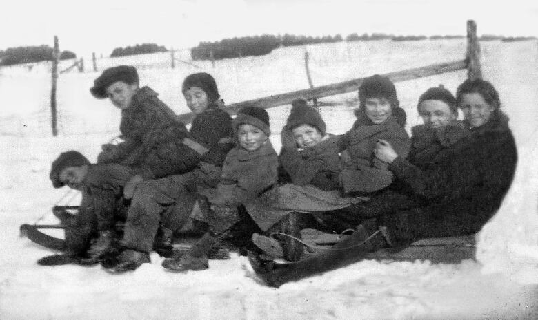 Black and white photo of 8 children all bundled up and piled onto a toboggan on a snowy hillside.