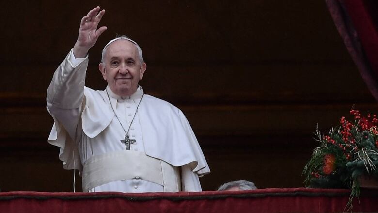 The Pope, wearing white, raises his right hand. Background is dark red.