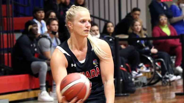 A woman with blond hair wearing a black-and-red sports uniform sits in a wheelchair holding a basketball in her left hand. 