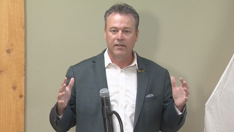 A man in a grey suit with a white shirt and no tie is standing in front of a wall gesturing with his arms wide open.