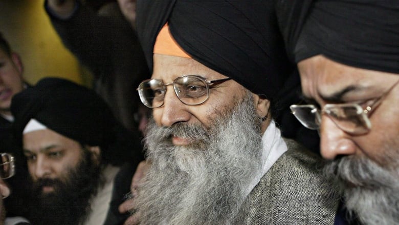 A man with a long grey beard and glasses is flanked by other men at a press scrum. He is wearing a black turban and a grey suit.