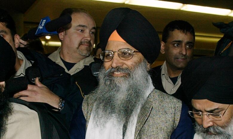 A man with a black turban and long grey beard is flanked by numerous people in a press scrum.