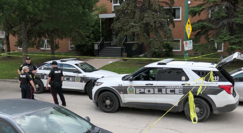 Three uniformed police officers and two marked police vehicles are seen on the street in front of a building, surrounded by yellow police tape.