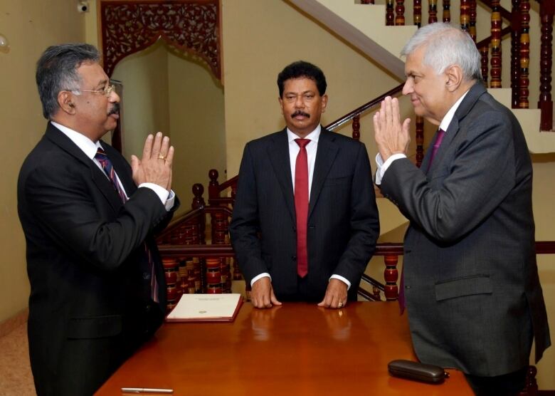 Three man stand around a table, with two of them folding their hands in a prayer-like position.