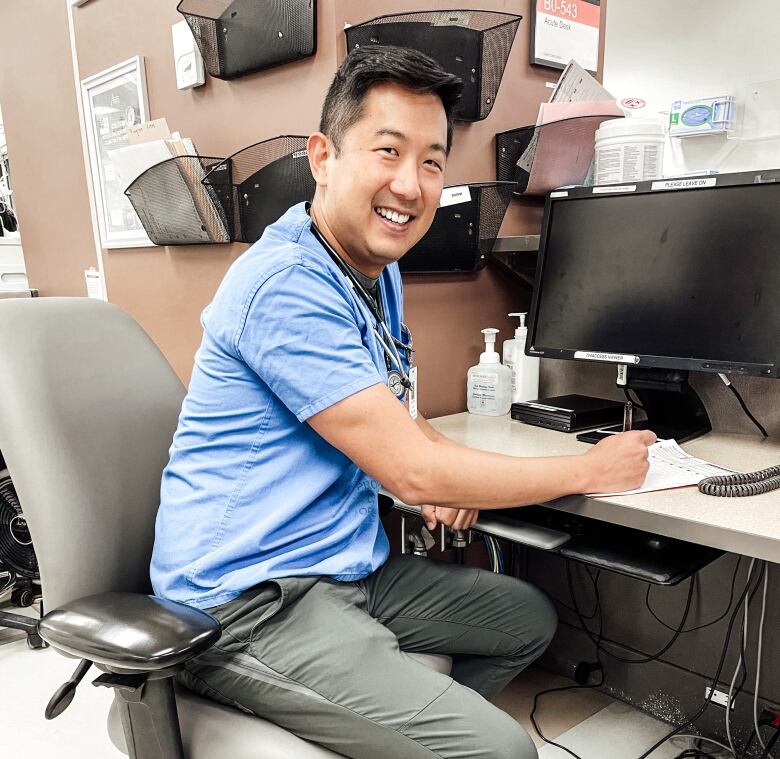 Dr. Justin Yan sitting at his work desk in London, Ontario, with a stethoscope around his neck. He is writing clinic notes. and smiling into the camera. 