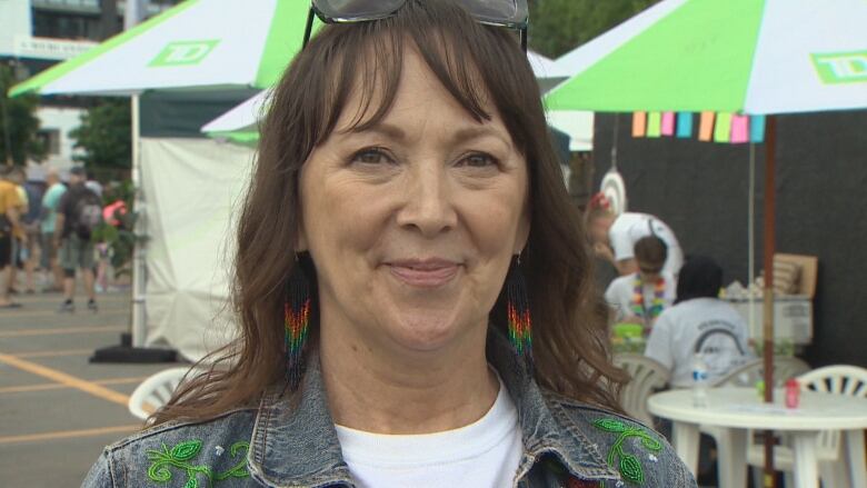 An adult woman outside. She has shoulder-length brown hair. She is wearing a denim jacket with a white t-shirt.