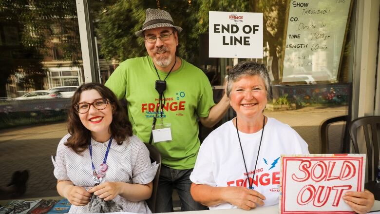 Three people sit at a table, two holding signs that read 'end of line' and 'sold out.'