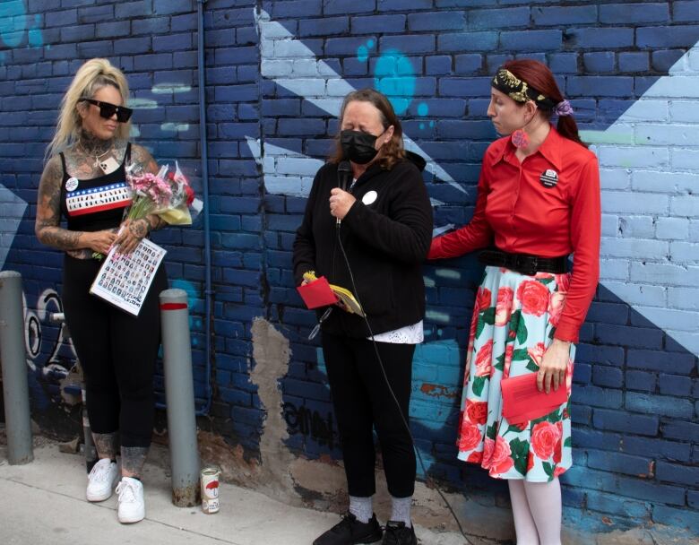 Three women stand in front of a blue wall. The woman in the center looks distressed and holds a microphone. The other women beside her are offering support. 