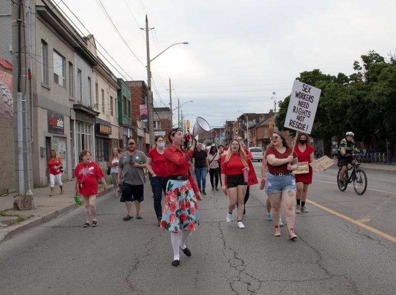 A crowd of people wearing read are led by a woman with a megaphone. One woman holds a sign that reads 'SEX WORKERS NEED RIGHTS NOT RESCUE'. 