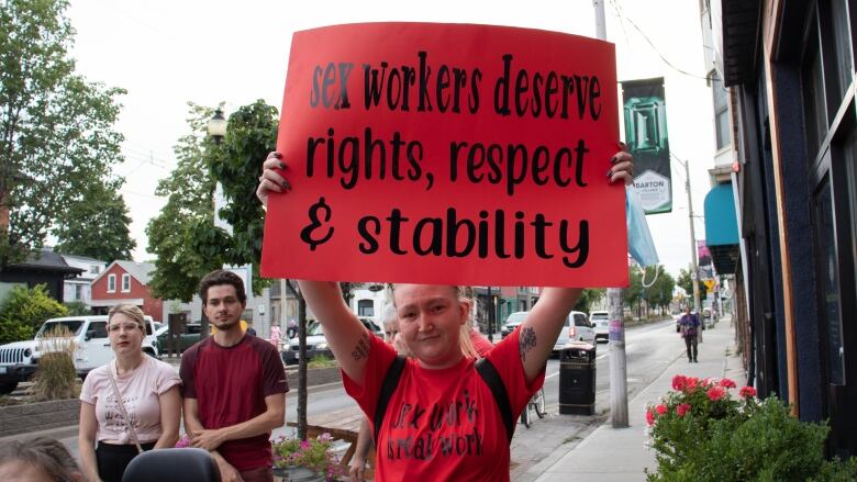 A woman in a red shirt that says 'SEX WORK IS REAL WORK' holds a red sign that says 'SEX WORKERS DESERVE RIGHTS, RESPECT AND STABILITY'